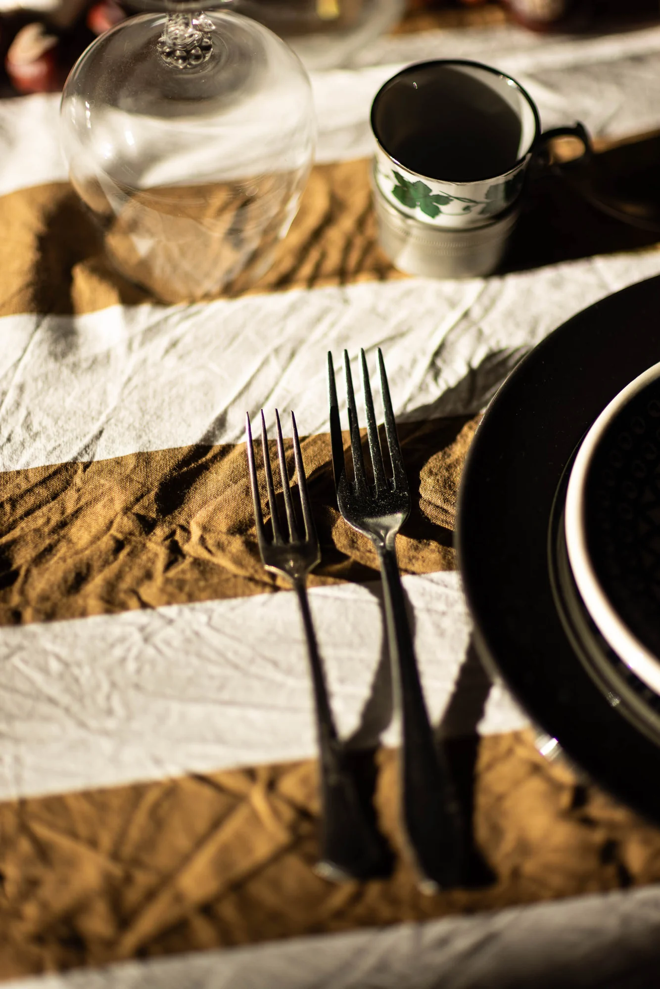 A place setting close up: ochre and white striped linen runner, an upturned wine glass, a cup with a green vine pattern, two old silver forks and a black plate