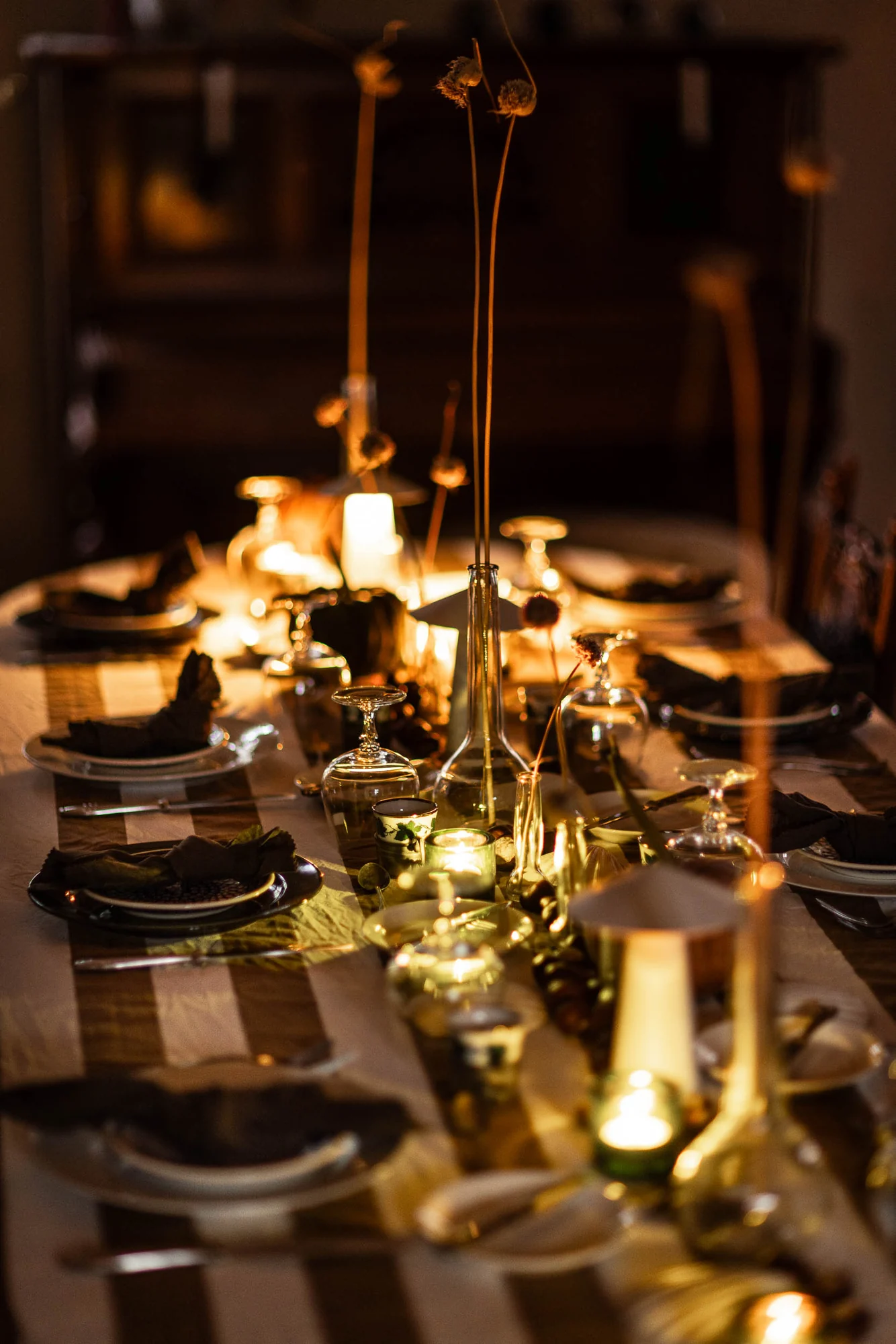 The long table seen end on by candlelight, upturned glasses, dark napkins and dried allium heads in a green bottle