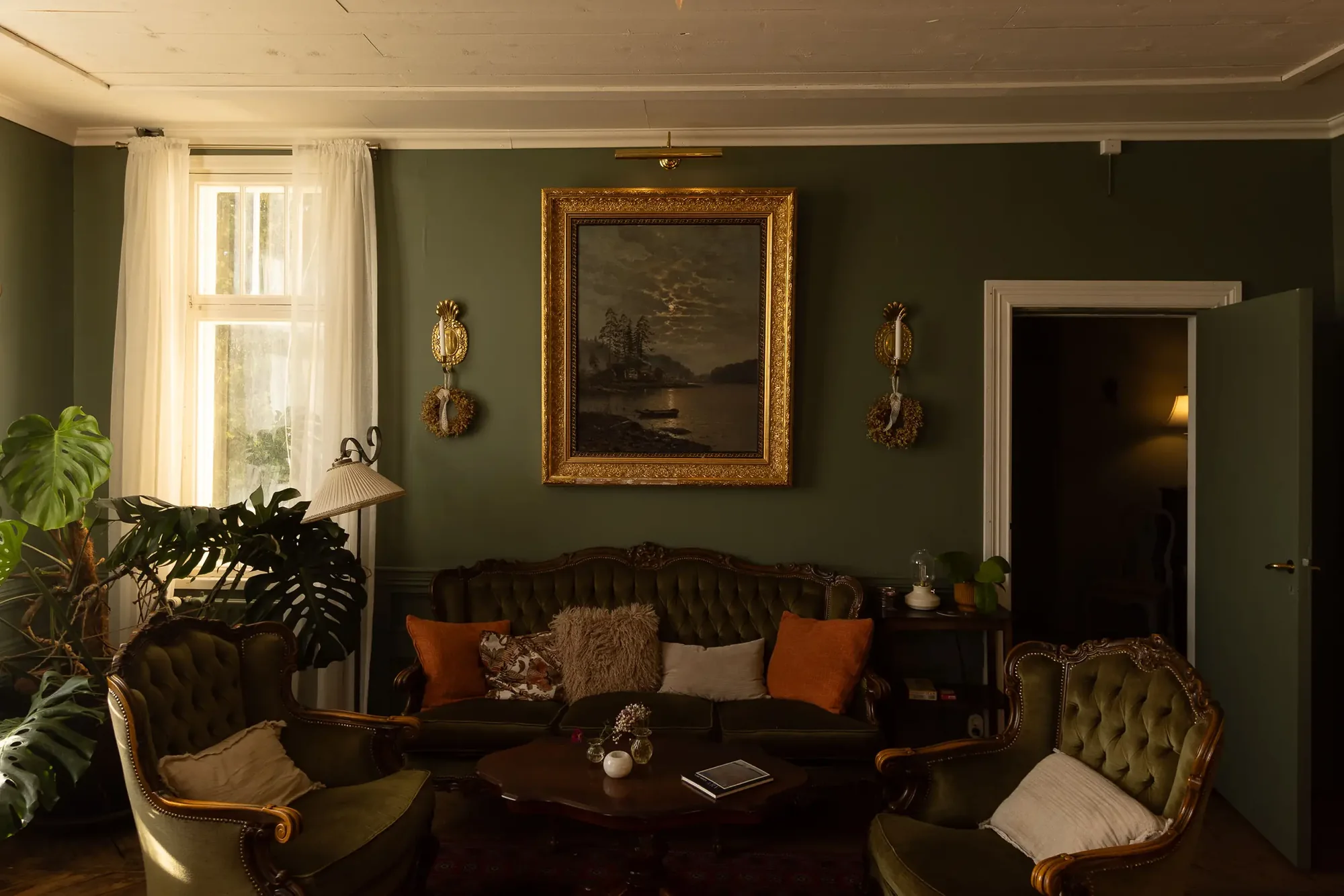 Salon in Kansjerf Gård mit dunkelgrünen Wänden, Samtsofas, goldgerahmtem Seestück und Monstera