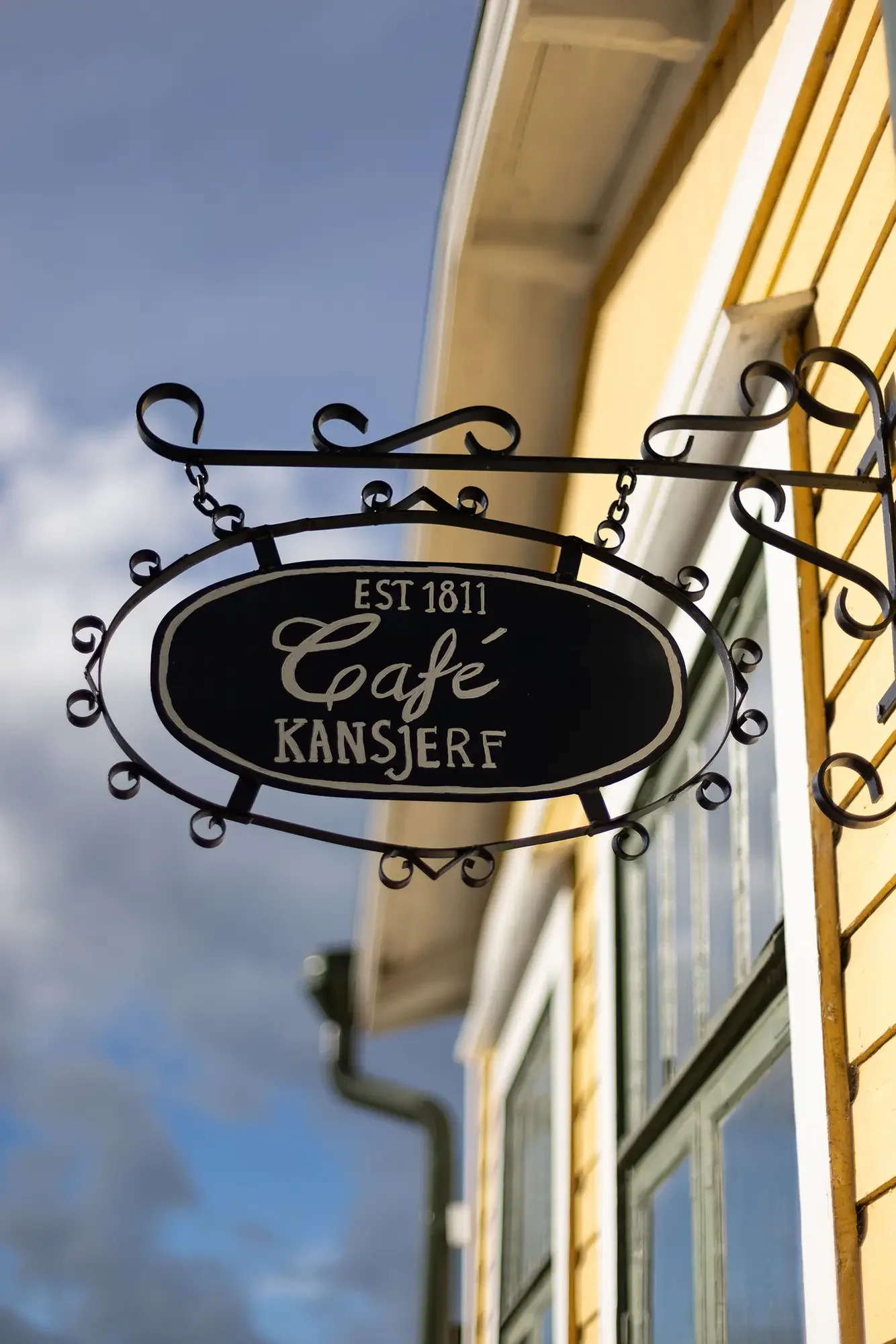 Schmiedeeisernes Schild Café Kansjerf, Est. 1811, an der gelben Fassade vor Wolkenhimmel